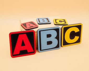 Może przedstawiać: Pięć drewnianych klocków z czarnymi obramowaniami i kolorowymi literami, które tworzą napis "ABC" w zabawnym wzorze. Klocki są ułożone w rzędzie na drewnianej powierzchni.