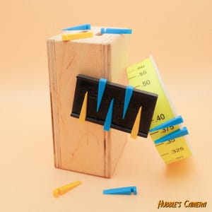 Op de afbeelding: Een zwart plastic gereedschap met gele en blauwe kunststof wiggen die eraan zijn bevestigd. Het gereedschap is gemarkeerd met "Guide + Align" en is bevestigd aan een houten doos. Een gele meetlint met zwarte markeringen is gedeeltelijk zichtbaar in de afbeelding. De meetlint geeft "0,30", "0,325", "0,35", "0,375", "0,40", "0,50", "0,55" en "0,60" aan.