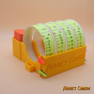 Puede incluir: Un dispositivo de plástico amarillo con cuatro carretes de cinta. La cinta está etiquetada con las letras A, B, C y E. El dispositivo está etiquetado con "Hubble's Camera".