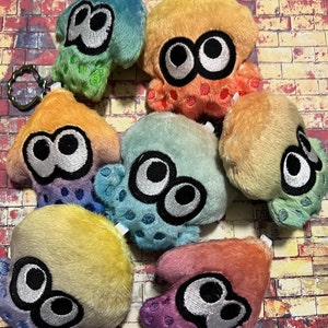 Puede incluir: Un conjunto de ocho peluches con forma de calamares en varios colores pastel. Cada calamar tiene ojos grandes y negros y una superficie texturizada y peluda.
