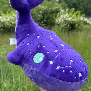 Könnte beinhalten: Ein tiefviolettes Wal-Stofftier mit aufgestickten hellblauen Sternbildern und Sternen auf dem Rücken. Der Wal hat eine weiche Textur, eine ausgeprägte Schwanzflosse und einen runden Aufnäher mit einem Mondmotiv an der Seite.