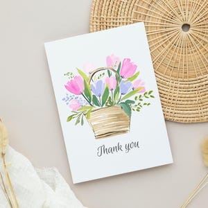 May include: A white greeting card with a watercolor illustration of a basket filled with pink and purple tulips and green foliage. The words "Thank you" are written below the illustration.