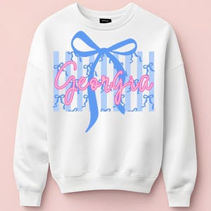 Op de afbeelding: Witte sweatshirt met een blauwe strik en de naam "Georgia" in roze script. De achtergrond heeft blauwe verticale strepen met kleine strikaccenten. Een casual top.