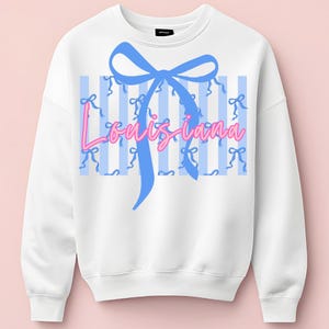 May include: White sweatshirt with a blue bow and the word "Louisiana" in pink script. The design includes blue vertical stripes and small pink bows, creating a preppy and feminine aesthetic.