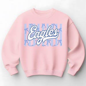 May include: A light pink sweatshirt with the word "Eagles" in a white and blue script font. The word is set against a background of light blue vertical stripes and small blue bows.