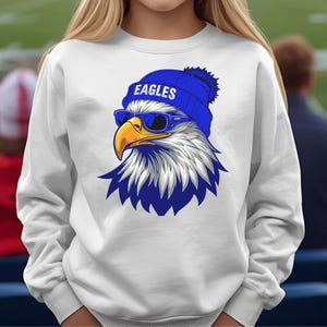 May include: White sweatshirt featuring a graphic of a stylized eagle head wearing a blue beanie with the word "EAGLES" and blue sunglasses. The eagle has a yellow beak and blue and white feathers.