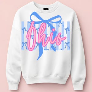 Könnte beinhalten: Weißes Sweatshirt mit dem Wort "Ohio" in rosa Schrift, akzentuiert durch eine blaue Schleife und blaue vertikale Streifen. Kleine blaue Schleifen sind über den Hintergrund verstreut. Ein lässiges, bequemes Oberteil.