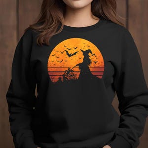 May include: Black sweatshirt with a Halloween-themed graphic. The design shows a witch silhouette on a bicycle against an orange sunset, with bats flying above. The graphic uses orange, black, and brown colours.