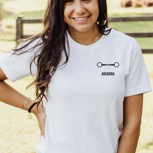 May include: A white t-shirt with a black equestrian bit design and the name "ARIANNA" printed on the front.