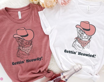 May include: Two t-shirts with a cat wearing a cowboy hat and the text "Gettin' Meowdy!" and "Gettin' Meowied!" on a white background.