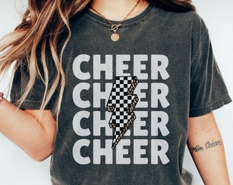 Pode incluir: Uma camiseta cinza com a palavra "CHEER" repetida quatro vezes em texto branco. Um gráfico de raio de xadrez preto e branco está centralizado na camiseta. A camiseta é de corte relaxado e é feita de 100% algodão.