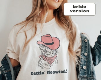 May include: Ivory relaxed fit t-shirt with a graphic of a cat wearing a pink cowboy hat and bandana. The text "Gettin' Meowied!" is printed on the shirt.  The shirt is made of 100% cotton.