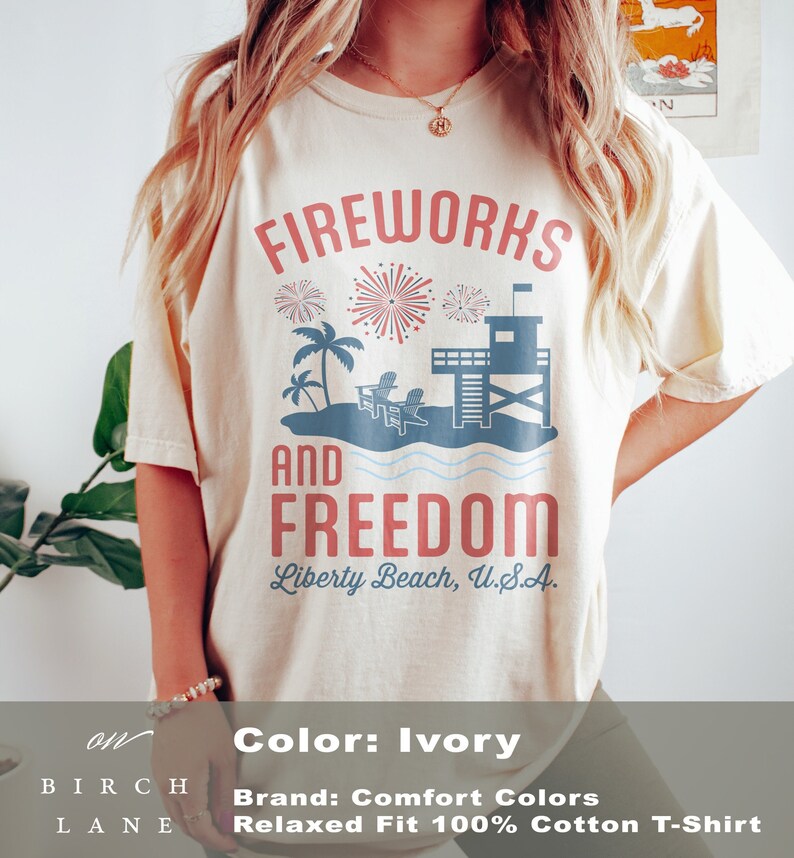 May include: Ivory-colored t-shirt with a graphic design featuring the words "Fireworks and Freedom" above a beach scene with a lifeguard tower, palm trees, and chairs. The text "Liberty Beach, U.S.A." is below.