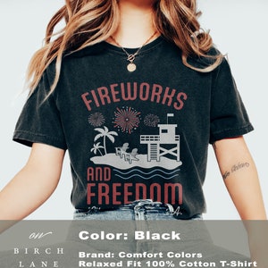 May include: Black t-shirt with a graphic design featuring the words "FIREWORKS AND FREEDOM". The design includes fireworks, palm trees, a lifeguard tower, and beach chairs. Made of 100% cotton.