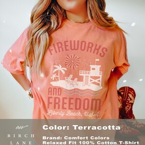 May include: Terracotta-colored t-shirt with the words "FIREWORKS AND FREEDOM" and a beach scene graphic. The graphic includes palm trees, chairs, and a lifeguard tower. The text "Liberty Beach, U.S.A." is also printed on the shirt.