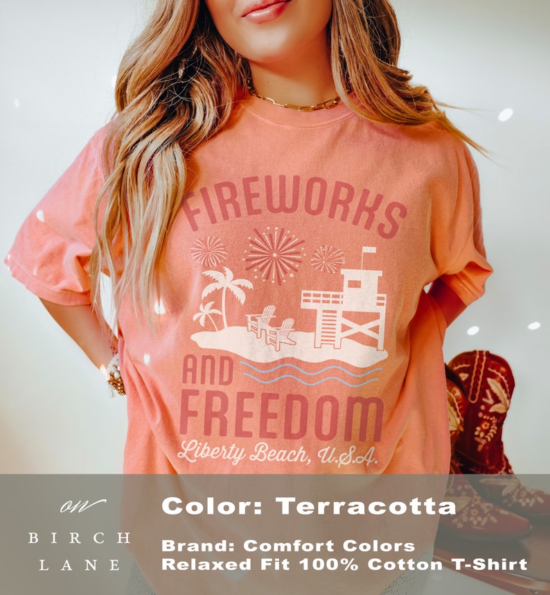 May include: Terracotta-colored t-shirt with the words "FIREWORKS AND FREEDOM" and a beach scene graphic. The graphic includes palm trees, chairs, and a lifeguard tower. The text "Liberty Beach, U.S.A." is also printed on the shirt.