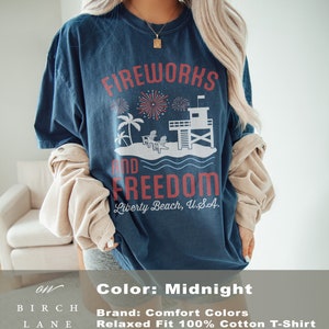 May include: Midnight blue t-shirt with a graphic that reads "Fireworks and Freedom" above a beach scene with a lifeguard tower and palm trees. The text "Liberty Beach, U.S.A." is below the graphic. The shirt is a relaxed fit.