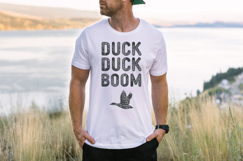 May include: White t-shirt with a black graphic print that says "Duck Duck Boom" and a silhouette of a duck flying.