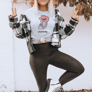 May include: A woman wearing a grey t-shirt with a cartoon cat wearing a cowboy hat and the text "meowdy". She is also wearing a black and white plaid jacket, black leggings, and white boots.