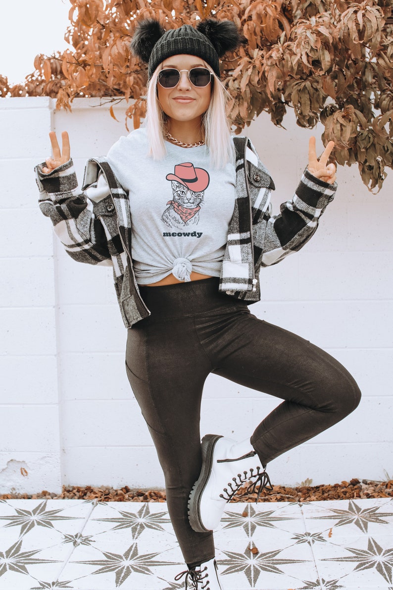 May include: A woman wearing a grey t-shirt with a cartoon cat wearing a cowboy hat and the text "meowdy". She is also wearing a black and white plaid jacket, black leggings, and white boots.
