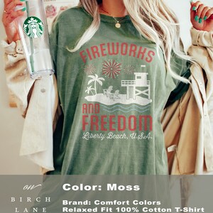 May include: Moss-colored, relaxed-fit cotton t-shirt with the words "FIREWORKS AND FREEDOM" in red. The design includes fireworks, palm trees, a lifeguard tower, and the text "Liberty Beach, U.S.A."