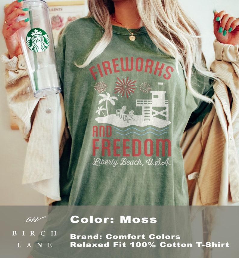 May include: Moss-colored, relaxed-fit cotton t-shirt with the words "FIREWORKS AND FREEDOM" in red. The design includes fireworks, palm trees, a lifeguard tower, and the text "Liberty Beach, U.S.A."