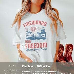 May include: White, relaxed-fit t-shirt with a graphic design. The design features the words "Fireworks and Freedom" above a beach scene with fireworks, palm trees, and a lifeguard tower. The text "Liberty Beach, U.S.A." is below.