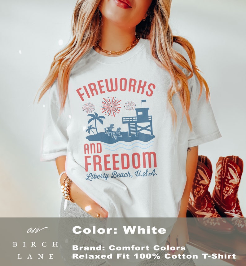 May include: White, relaxed-fit t-shirt with a graphic design. The design features the words "Fireworks and Freedom" above a beach scene with fireworks, palm trees, and a lifeguard tower. The text "Liberty Beach, U.S.A." is below.