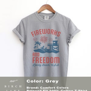 May include: A grey, relaxed-fit, 100% cotton t-shirt with a graphic design. The design features the words "FIREWORKS AND FREEDOM" in red, with a beach scene including fireworks, palm trees, and a lifeguard tower. The text "Liberty Beach, U.S.A." is below.
