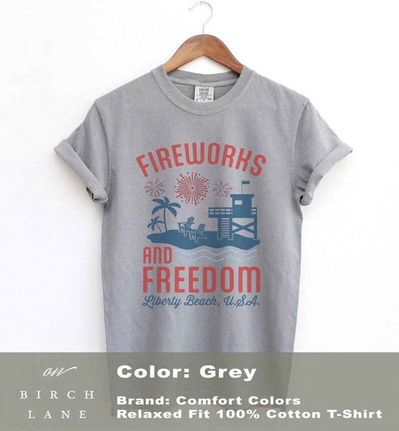 May include: A grey, relaxed-fit, 100% cotton t-shirt with a graphic design. The design features the words "FIREWORKS AND FREEDOM" in red, with a beach scene including fireworks, palm trees, and a lifeguard tower. The text "Liberty Beach, U.S.A." is below.