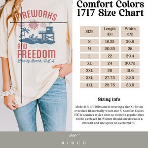 May include: Cream-colored t-shirt with a graphic design featuring fireworks, palm trees, a lifeguard tower, and the text "Fireworks and Freedom, Liberty Beach, U.S.A." A size chart is also visible.