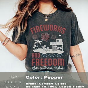 May include: Pepper-colored t-shirt with a graphic design featuring fireworks, palm trees, a lifeguard tower, and the words "Fireworks and Freedom, Liberty Beach, U.S.A." The shirt is a relaxed fit and made of 100% cotton.