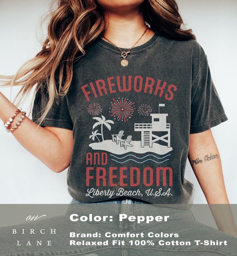 May include: Pepper-colored t-shirt with a graphic design featuring fireworks, palm trees, a lifeguard tower, and the words "Fireworks and Freedom, Liberty Beach, U.S.A." The shirt is a relaxed fit and made of 100% cotton.