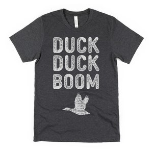 May include: A dark grey t-shirt with a white distressed graphic that says "Duck Duck Boom" and a white silhouette of a duck flying above the text.