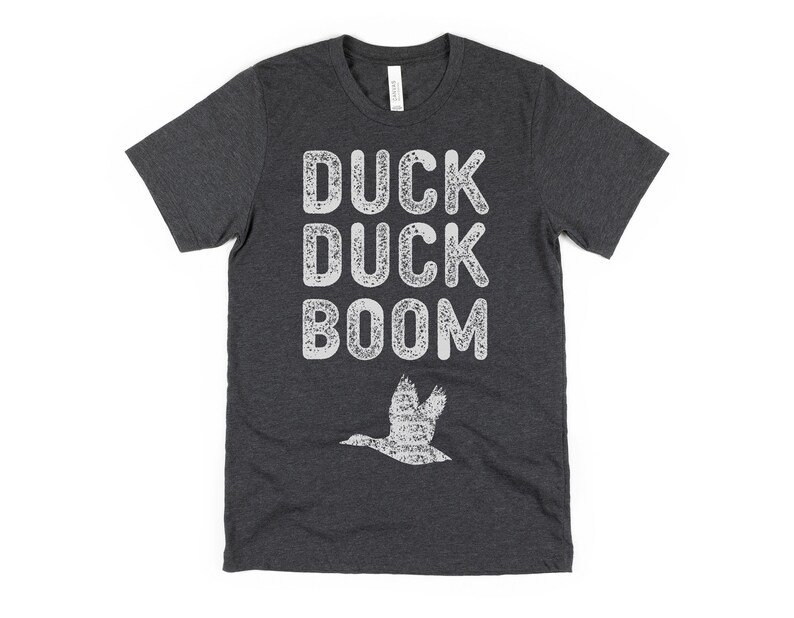 May include: A dark grey t-shirt with a white distressed graphic that says "Duck Duck Boom" and a white silhouette of a duck flying above the text.
