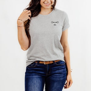 May include: A gray t-shirt with a black equestrian bit and the letters "ECL" printed on the front. The shirt is being worn by a person with long brown hair and a brown leather belt.