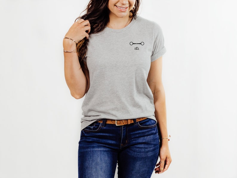 May include: A gray t-shirt with a black equestrian bit and the letters "ECL" printed on the front. The shirt is being worn by a person with long brown hair and a brown leather belt.