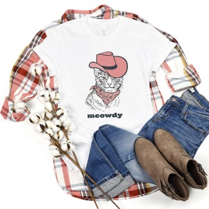 May include: White t-shirt with a cartoon illustration of a cat wearing a cowboy hat and bandana. The text "meowdy" is printed below the cat.
