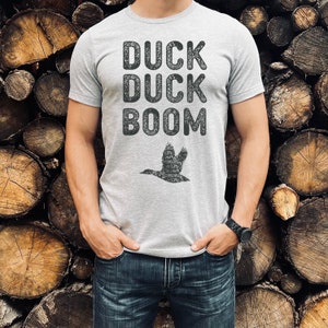 May include: A grey t-shirt with the text "Duck Duck Boom" and a silhouette of a flying duck. The shirt is being worn by a person standing in front of a stack of wooden logs.