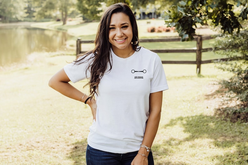 May include: A white t-shirt with a black equestrian bit design and the name "ARIANNA" printed on the front.
