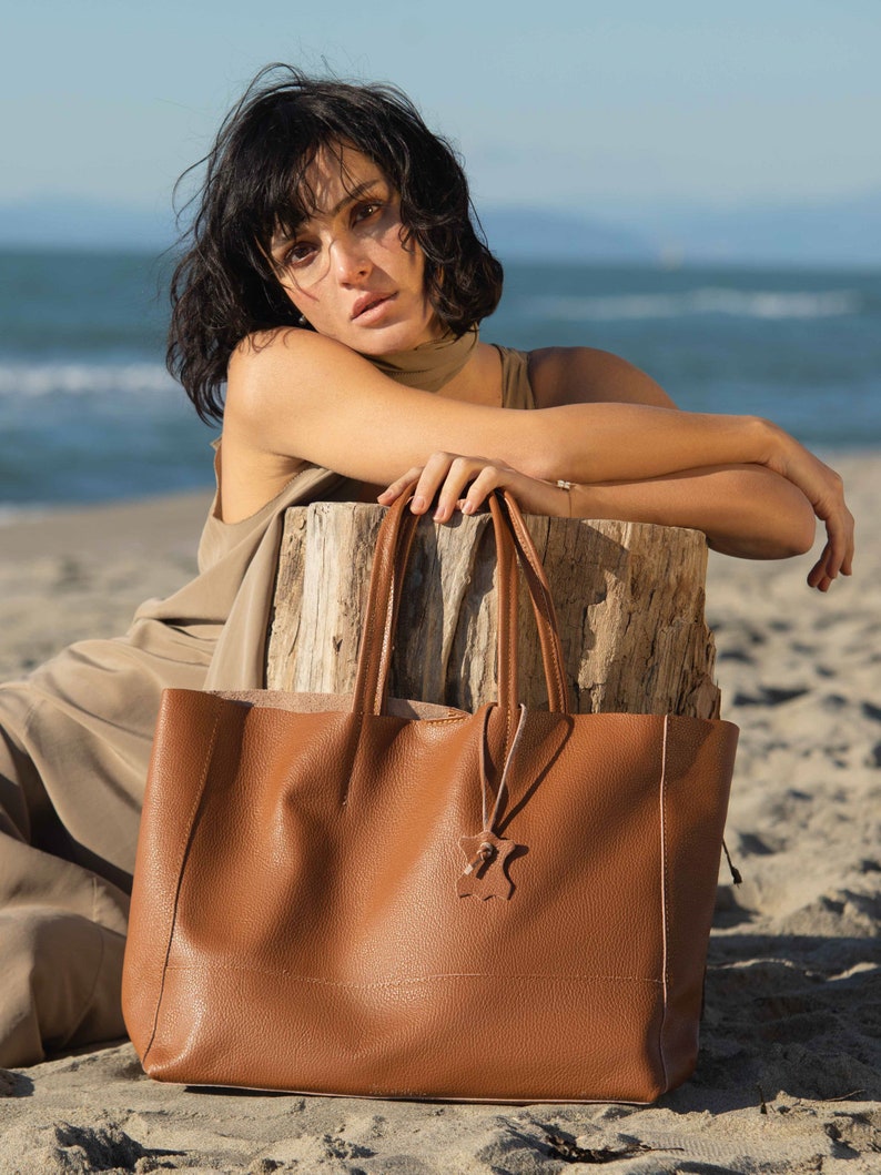 a woman sitting on the beach with a brown bag