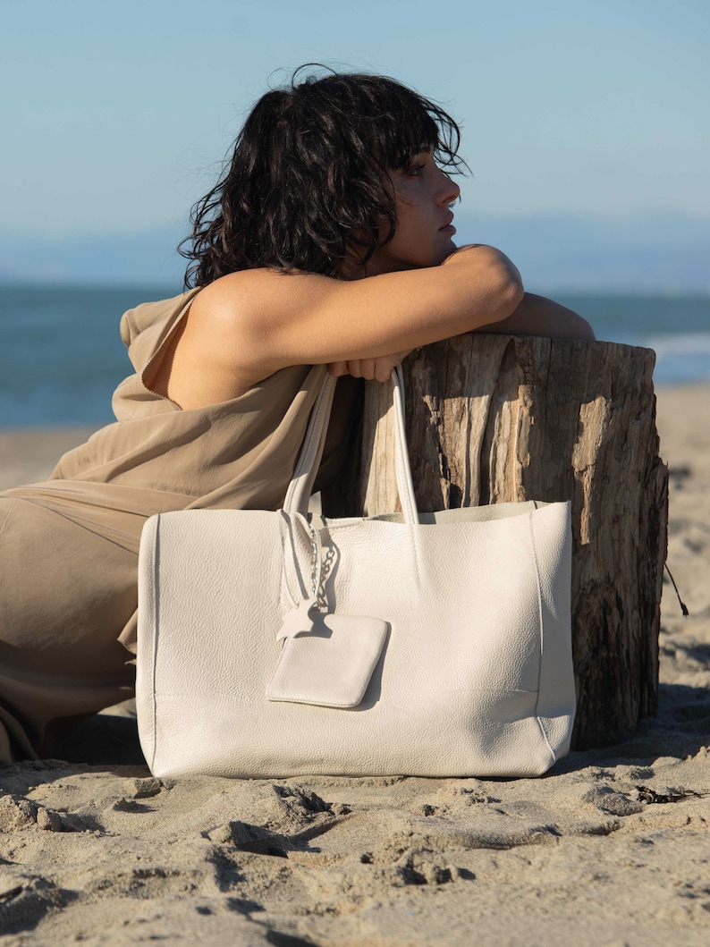 a woman sitting on the beach with a white bag
