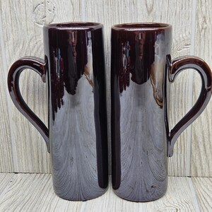 May include: Two tall, cylindrical mugs with a glossy, dark brown finish. Each mug features a smooth, reflective surface and a curved handle attached to the side. The mugs have a uniform, straight-sided design with a simple, open top rim.
