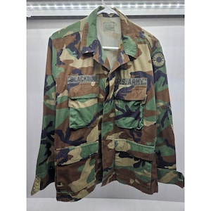 May include: Camouflage pattern military jacket with multiple pockets and the name "BLACKBURN" above the left chest pocket. The right chest pocket has "U.S. ARMY" printed on it. The jacket is green, brown, and black.