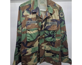 Puede incluir: Chaqueta militar con estampado de camuflaje con múltiples bolsillos. El nombre "BLACKBURN" está encima del bolsillo izquierdo del pecho y "U.S. ARMY" está a la derecha. La chaqueta es verde, marrón y negra.
