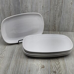 May include: A set of four white rectangular ceramic plates with rounded corners. One plate is positioned vertically on a small black stand, while the other three plates are stacked horizontally in front of it, creating a clean and uniform appearance.