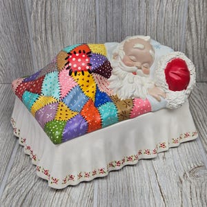 May include: A ceramic figurine of a sleeping Santa Claus tucked under a colorful patchwork quilt. The quilt features various patterns including polka dots and stars in red, yellow, blue, purple, and green. Santa wears a red hat with white trim.