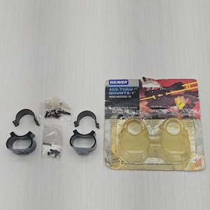 May include: A set of black metal Weaver see-thru mounts for a Winchester 70 rifle, including two pairs of rings and small screws in plastic bags, alongside the original cardboard packaging with a clear plastic blister pack and printed text.