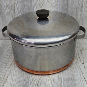 May include: A stainless steel stockpot with a matching lid and a dark brown knob handle. The pot features a wide, cylindrical body with two side handles and a distinct copper-colored base, providing a clear view of the metallic construction.