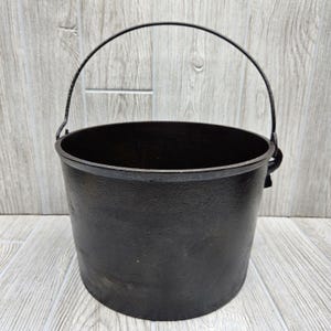 May include: A black cast iron pot with a metal handle. The pot is empty and has a smooth, slightly worn surface.